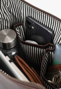 Gestreifter Stoffbeutel, der ein schwarzes Smartphone, Schlüssel, eine Ledergeldbörse, eine Wasserflasche, ein Notizbuch und eine Tube Creme enthält.