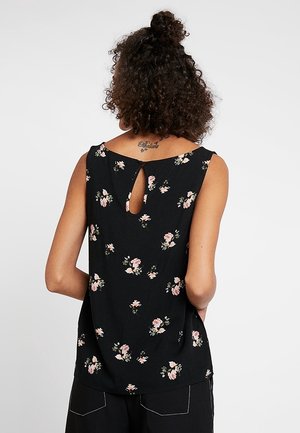 Vrouw met krullend haar die een mouwloos zwart topje draagt met een roze bloemenprint en een kleine sleutelgat uitsnijding bovenaan de rug.