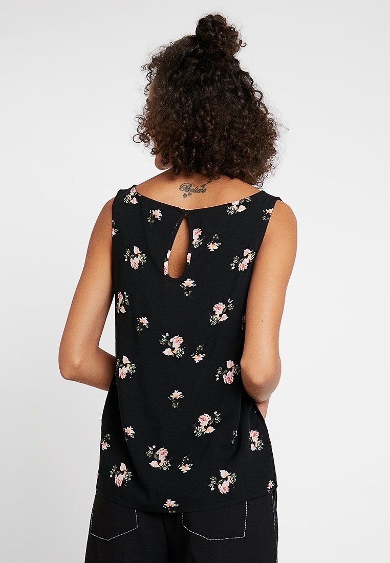 Femme aux cheveux bouclés portant un haut noir sans manches avec un imprimé floral rose et une petite découpe en forme de goutte au niveau du haut du dos.