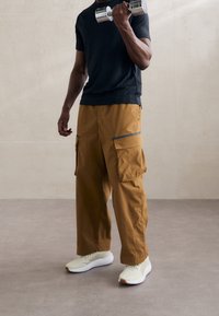 Kargo kalhoty hnědé barvy z širokého materiálu, s bočními kapsami a šedými akcenty. Černé tričko a světlé tenisky doplňují celé oblečení.