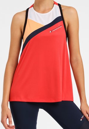 Vrouw draagt een mouwloos sporttopje in rood, perzik, marineblauw en wit met zwarte bandjes, gecombineerd met marineblauwe legging, staat met ontspannen handen.