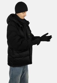 Schwarze Daunenjacke mit Kapuze, gestrickte schwarze Mütze, Schal und Handschuhe. Weicher Stoff, glatte Textur, mit einem Logodetail auf der Jacke.