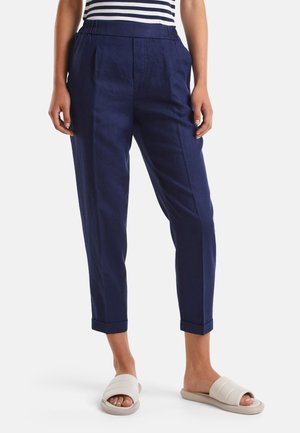 Kobieta w granatowych, skróconych spodniach z mankietami, w paskowanym topie i białych sandałach na płaskiej podeszwie stoi na białym tle.