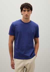 Junger Mann mit dunklem lockigem Haar, der ein schlichtes blaues Kurzarm-T-Shirt und helle beiges Hosen trägt, vor einem hellgrauen Hintergrund.