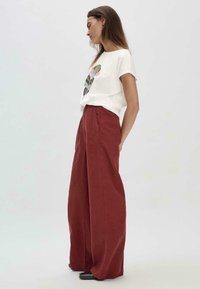 Witte katoenen T-shirt met een kleurrijk cactusontwerp, gecombineerd met hoogsluitende, wijde roestkleurige broeken. Zwarte schoenen.