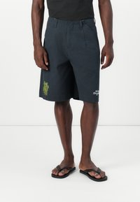 Donkergrijze shorts van lichtgewicht stof, met een geborduurd groen ontwerp aan de zijkant en een relaxte pasvorm. Gecombineerd met zwarte slippers.
