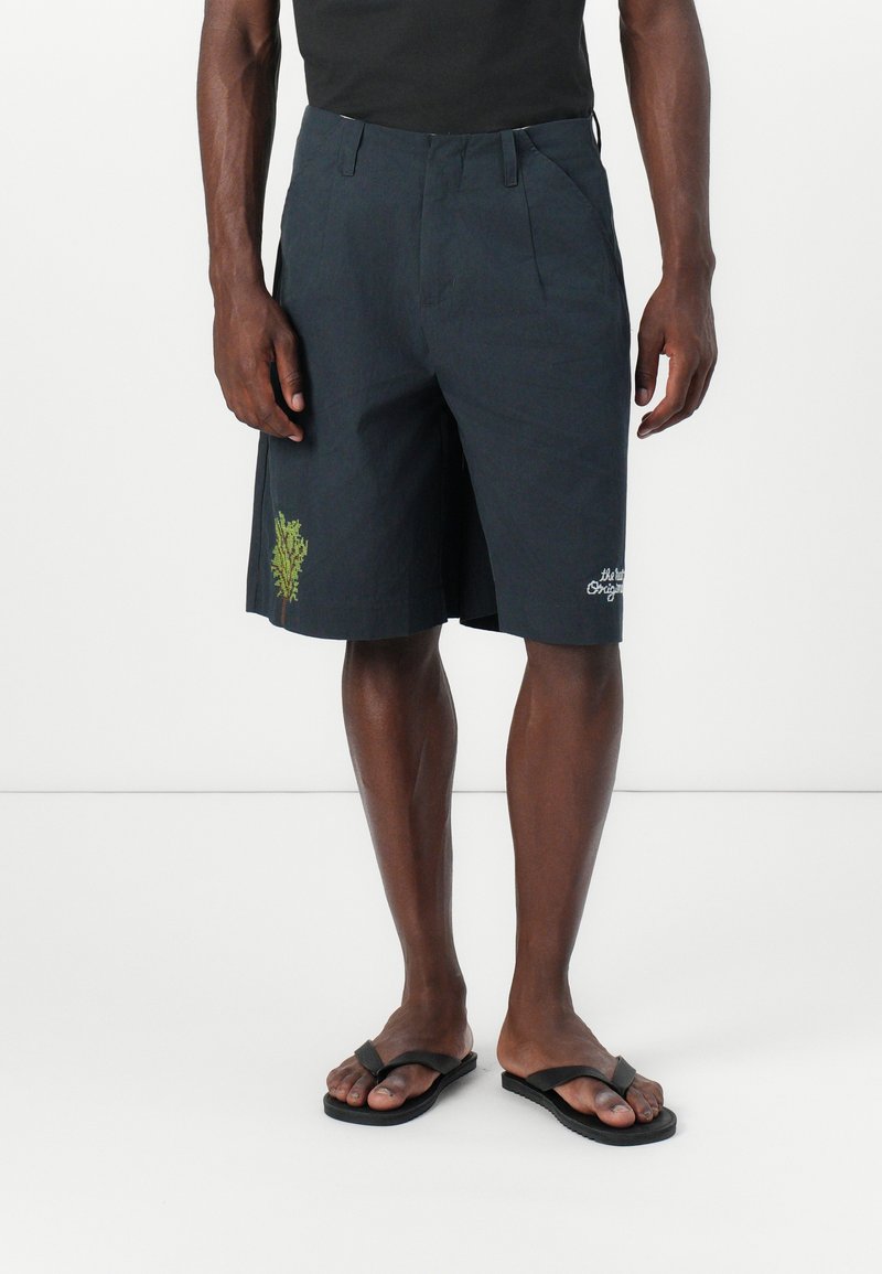 Donkergrijze shorts van lichtgewicht stof, met een geborduurd groen ontwerp aan de zijkant en een relaxte pasvorm. Gecombineerd met zwarte slippers.