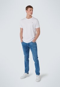 Witte crew-neck t-shirt met een klein logo, gecombineerd met blauwe slim-fit jeans en witte sneakers, staand tegen een lichtgrijze achtergrond.