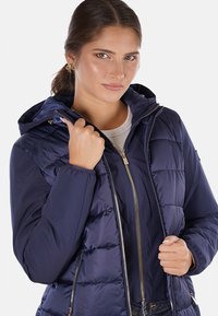 Giacca puffer blu navy con cappuccio removibile, chiusura frontale con zip e polsini a coste; presenta dettagli in oro e texture trapuntata.