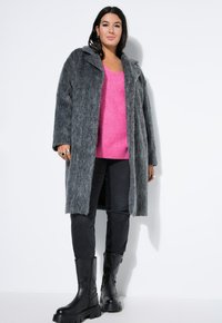 Abrigo de piel sintética gris con cuello de muesca, llevado sobre un suéter de punto rosa brillante. Jeans skiny negros y botas de cuero negras gruesas completan el look.