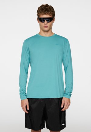 Lyseblå langærmet skjorte med en glat tekstur og et logo på brystet, parret med sorte shorts med en subtil glans.