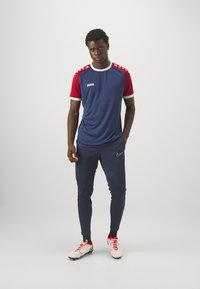 Completo sportivo composto da una maglia blu navy con maniche corte rosse e dettagli bianchi, abbinata a pantaloni attillati blu navy e scarpe bianche con dettagli rossi.