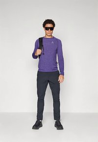 Homem a usar óculos de sol pretos, camiseta de manga longa desportiva roxa, calças pretas, sapatilhas pretas, segurando a alça de uma mochila preta, em fundo branco.