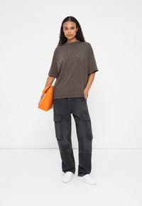 Kvinde står i løse mørkegrå cargobukser, oversized falmet sort t-shirt, hvide sneakers og holder en orange clutch-taske mod en hvid baggrund.