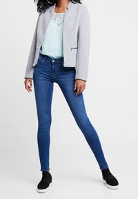 Grau gestreifte Blazer über einer hellblauen Bluse mit Spitzenakzenten, kombiniert mit schmalen blauen Jeans und schwarzen Slippern.
