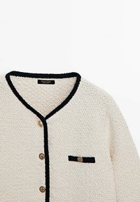Crème gebreide cardigan met zwarte afwerking, drie decoratieve gouden knopen aan de voorkant, en een kleine zwart-afgeborde pocket met gouden knop op de borst.