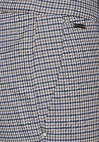 Tkanina s tkaným houndstooth vzorem v modré, hnědé a krémové barvě. Obsahuje boční kapsu a detail loga na pase.