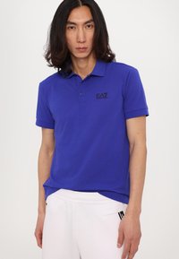 Royalblaue Poloshirt aus Baumwolle mit Kragen, kurzen Ärmeln und einem schwarzen Logo auf der linken Brust. Trägt eine weiße Hose.
