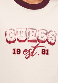 Λευκό μπλουζάκι με μπορντό λεπτομέρειες και υφασμάτινο γραφικό. Διαθέτει τολμηρό "GUESS" σε κόκκινο με λάμψεις, τον αριθμό "19" και το "est. 81" σε καλλιγραφία.