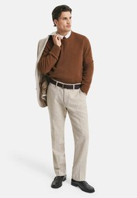 Mann trägt einen braunen Pullover, beigen Hosen und schwarze Schuhe, hält einen hellbeigen Mantel über der Schulter und steht vor einem weißen Hintergrund.