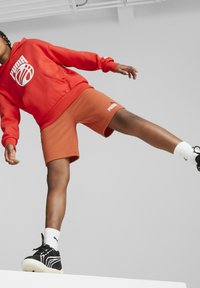 Červená mikina s bílým logem, v kombinaci s oranžovými šortkami. Černé tenisky, bílé ponožky. Důraz na ležérní, sportovní design a živé barvy.