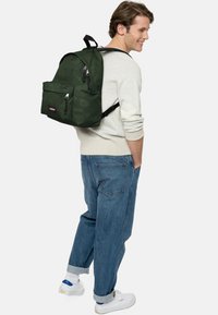Mochila de camuflaje verde con un bolsillo delantero, cierre de cremallera y correas ajustables, llevada sobre un suéter gris y unos vaqueros azules holgados.