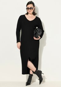 Schwarzes, geripptes Midikleid mit V-Ausschnitt und langen Ärmeln, das einen seitlichen Schlitz aufweist. Kombiniert mit spitz zulaufenden, schwarzen Stiefeletten und einer schwarzen Clutch.