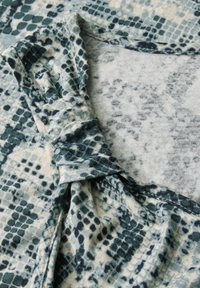 Zielono-kremowa tkanina w wzór węża z wywinięciem przy dekolcie; miękka tekstura, zblendowane kolory i brak widocznych elementów metalowych.