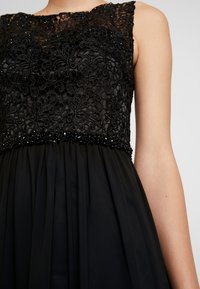 Svart klänning med en spetslivstycke med blommönster, dekorerad med pärlor, och en flödande chiffongkjol. Ärmlös design.