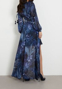 Robe longue et transparente de couleur bleu foncé avec des motifs floraux. Présente des manches bouffantes, une ceinture à la taille et une fente sur le côté. Assortie avec des talons aiguilles.