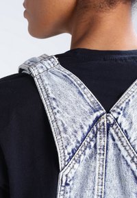 Närbild på en persons axel som bär syra-tvättade denimoverall med justerbara metallspännen över en svart skjorta.