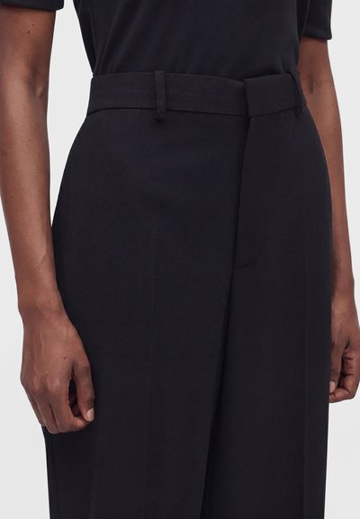 Κοντινό πλάνο ενός ατόμου με μαύρο tailored παντελόνι ψηλής μέσης με θηλιές για ζώνη και μαύρο τοπ, με τα χέρια να ακουμπούν στα πλευρά του.