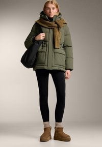 Olívazöld steppelt kabát kapucnival és két első zsebbel, fekete leggings-szel, világosbarna bokacsizmával és világosbarna sállal párosítva. Fekete válltáska.