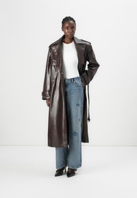Glänzender brauner Leder-Trenchcoat mit beltem Bund, getragen über einem weißen Oberteil und ausgewaschenen blauen weiten Jeans, kombiniert mit schwarzen spitzen Absätzen.