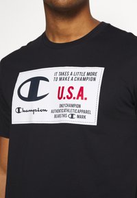 Černé tričko s bílou nášivkou, na které je logo značky Champion a text: "Chce to ještě něco navíc, abychom se stali šampiony USA." v červených a černých písmenech.