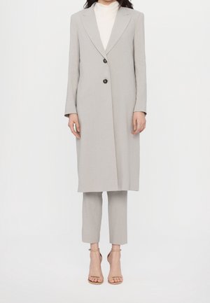 Femme portant un long blazer gris clair sur un pantalon court assorti et des talons ouverts couleur nude, debout sur un fond blanc.