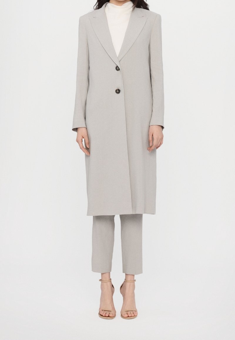 Femme portant un long blazer gris clair sur un pantalon court assorti et des talons ouverts couleur nude, debout sur un fond blanc.