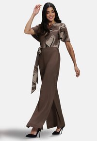 Brown jumpsuit with a flowing design, featuring a patterned top, wide legs, and a waist tie. Made of soft fabric, detailed with abstract prints.