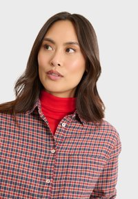 Chemise à carreaux rouge et marine avec un design à boutons, dotée d'une poche et de boutons blancs, portée par-dessus un col roulé rouge vif.