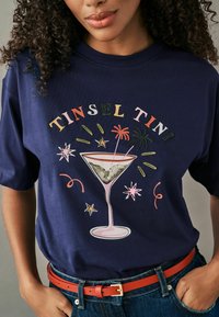 Marineblå t-shirt med et grafisk design af et martini glas med farverig tekst "Tinsel Tini" og livlige dekorationer. Lavet af bomuldsmateriale, afslappet pasform.