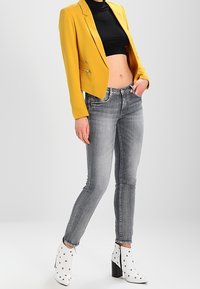 Osoba v zkrácené hořčicové blejzrové saku, černém crop topu, šedých skinny džínách a bílých kotníkových botách se černými nýty, pózuje s jednou rukou v kapse.