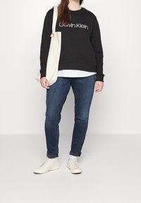Černá mikina Calvin Klein s bílým logem, vrstvená přes bílou košili. Nošena s modrými džíny a béžovými šněrovacími teniskami. Krémová taška.