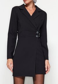 Femme portant une robe blazer noire ajustée avec deux boutons sur les côtés, des manches longues et des collants noirs translucides, devant un fond clair uni.