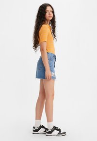 Jonge vrouw met lang, krullend haar staat opzij en draagt een oranje gestreepte korte mouwen top, een denimmuts, witte sokken en zwart-witte sneakers.