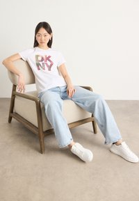 Weißes T-Shirt mit mehrfarbigem "DKNY"-Grafik, hellblaue Jeans und weiße Sneakers. Modell sitzt auf einem Holzstuhl vor einem neutralen Hintergrund.