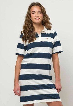 Młoda kobieta z długimi kręconymi włosami, ubrana w krótką sukienkę polo w niebiesko-białe poziome pasy, stojąca na tle jednolitego tła.