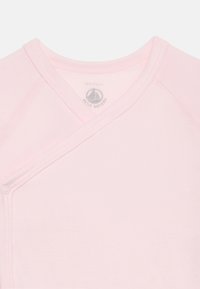 Chemise d'emmaillotage pour bébé de couleur rose doux avec fermeture à pression sur le côté, arborant un logo discret de voilier à l'intérieur du col, taille 12 mois.