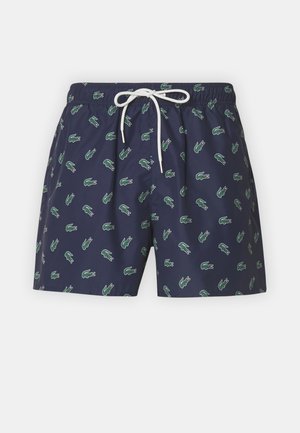 Tengerészkék úszónadrág könnyű anyagból, ismétlődő zöld krokodilmintával és fehér húzózsinórral a deréknál.