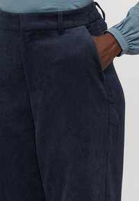 Navy corduroy trousers with vertical ribbing, featuring side pockets and a relaxed fit. The waistband has a subtle elastic detailing.