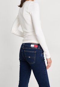 Jeans de mezclilla azul oscuro de corte ajustado, con un parche visible del logo de Tommy Jeans en el bolsillo trasero, combinados con una blusa blanca de manga larga ceñida.